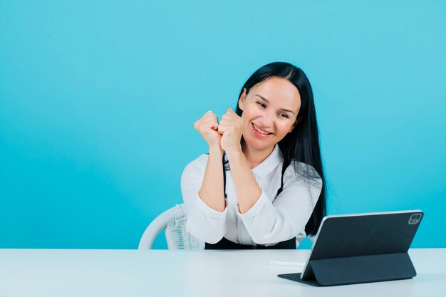 Happy blogger girl is holding hands together by posing to tablet camera on blue background