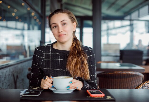 Happy blogger girl is looking at camera by holding coffee cup in cafe