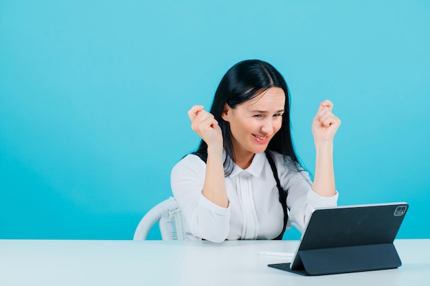 Happy blogger girl is raising up her fists by looking at tablet camera on blue background