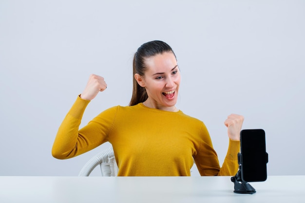 Happy blogger girl is raising up her fists in front of mobile camera on white background