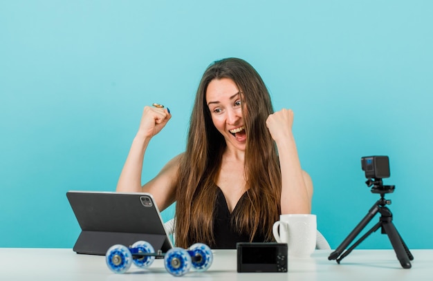 Happy blogger is posing at tablet camera by raising up fists on blue background