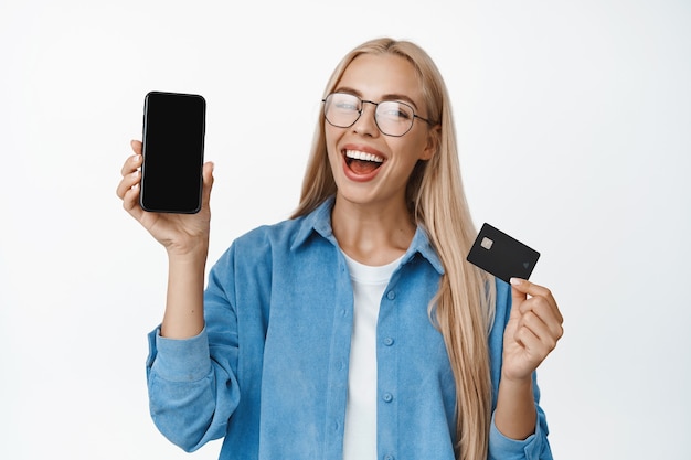 Happy blond female model in glasses, smiling and laughing, showing mobile phone screen and credit card, recommending smartphone interface on white.