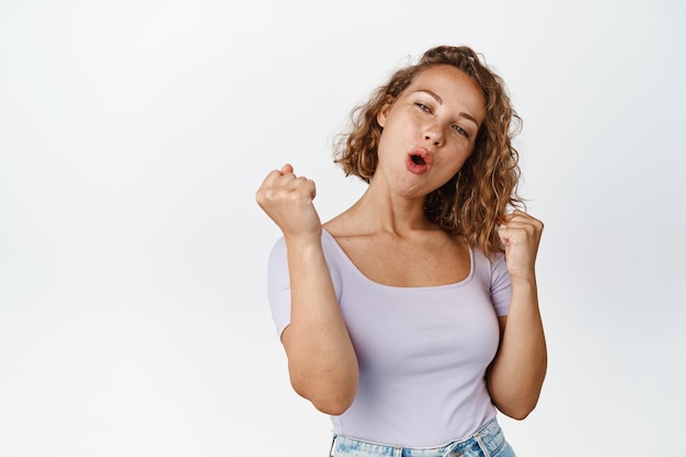 Happy blond girl celebrating victory, shouting and triumphing, winning prize, standing in tshirt over white background.