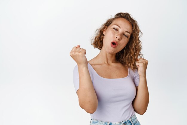 勝利を祝う幸せなブロンドの女の子、叫び、勝利、賞を獲得、白い背景の上にTシャツで立っています。