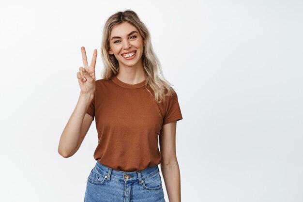 Happy blond girl shows peace, v-sign gesture and smiling, standing near empty copy space for advertisement on white studio