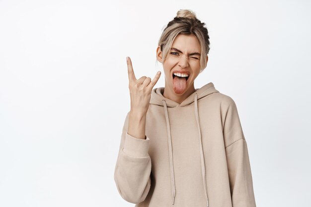 Happy blond girl shows tongue and heavy metal horns enjoying something having fun standing in beige hoodie against white background