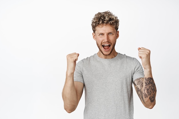 Happy blond guy triumphing sreaming with joy and shaking fists celebrating achieve success standing over white background in grey casual tshirt