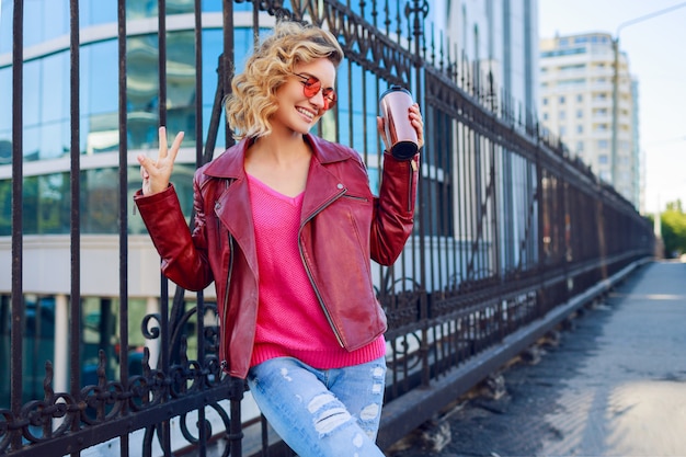 Счастливая белокурая женщина представляя на современных улицах, выпивая кофе или капучино. Стильный осенний наряд, кожаная куртка и вязаный свитер. Розовые очки.