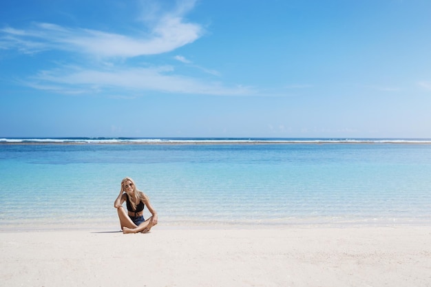 Счастливая блондинка сидит, скрестив ноги, песчаный пляж, приморский берег, наслаждается отпуском, летним путешествием, Мальдивами, загорает возле голубого спокойного моря, вид на океан, расслабляющий райский остров, курорт