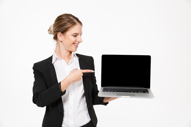 Happy blonde business woman showing blank laptop computer screen while looking and pointing at him over white wall