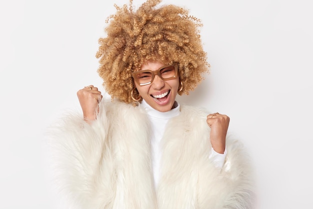 Foto gratuita felice bionda giovane donna dai capelli ricci scuote i pugni celebra il successo fa sì gesto accetta grande offerta indossa occhiali e pelliccia bianca pone indoor raggiunge l'obiettivo sì siamo campioni