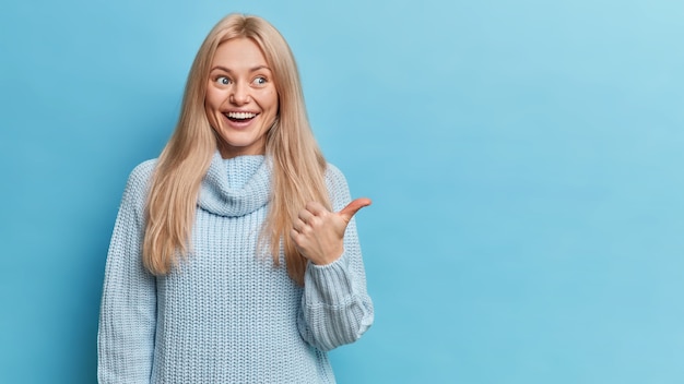 happy blonde European woman has glad expression dressed in knitted sweater points thumb at copy space