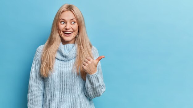happy blonde European woman has glad expression dressed in knitted sweater points thumb at copy space