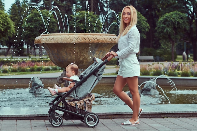 Happy blonde female dressed in a white blouse and shorts walking with baby stroller near a fountain in the city park.