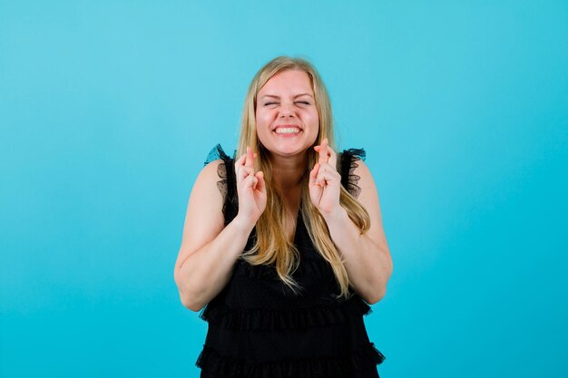 Happy blonde girl is crossing fingers on blue background