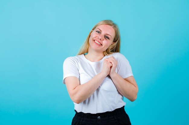 Happy blonde girl is looking away by holding hands together on chest on blue background