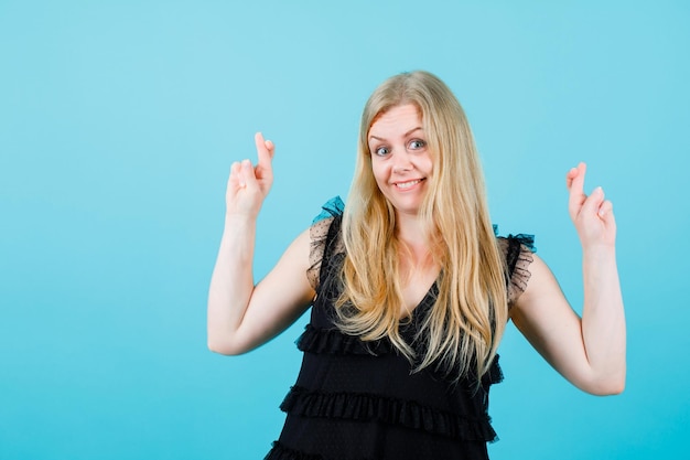 Free photo happy blonde girl is looking at camera by crossing fingers on blue background