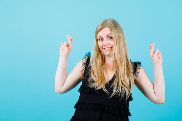 Happy blonde girl is looking at camera by crossing fingers on blue background