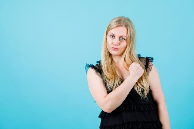 Happy blonde girl is looking at camera by holding fist near shoulder on blue background