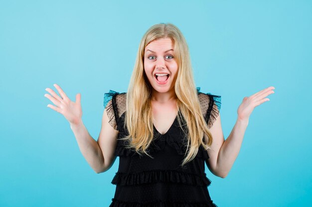 Happy blonde girl is looking at camera by opening wide her hands on blue background