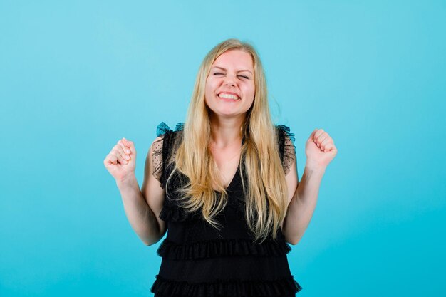 Happy blonde girl is raising up her fisst on blue background