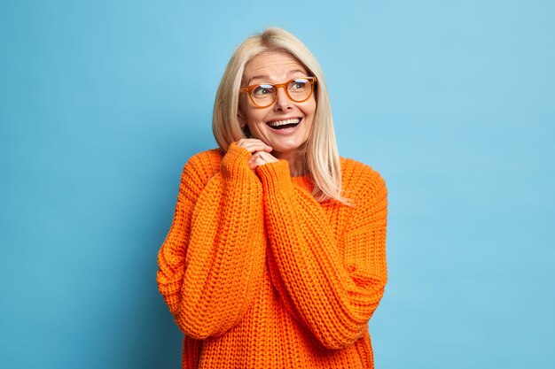 Счастливая белокурая старшая европейка вспоминает что-то приятное, радостно смеется и держит руки вместе, носит оптические очки, свободный вязаный оранжевый джемпер.