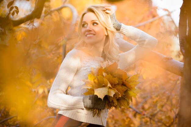 カエデの葉の束で立って、屋外で写真家のためにポーズをとっているきれいな女性を振り返って幸せなブロンド