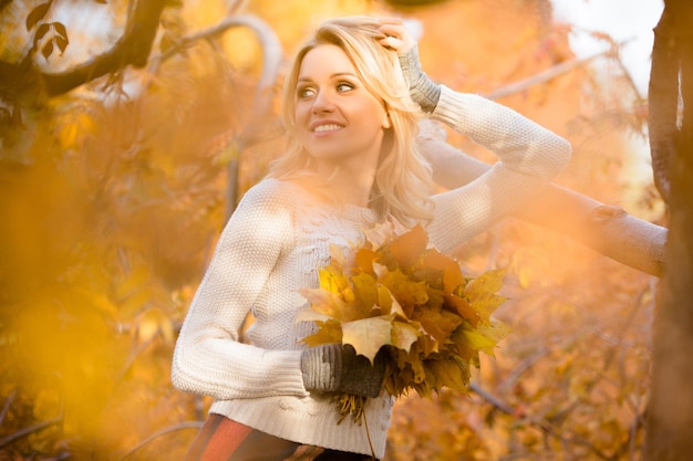 Счастливая блондинка, стоящая с кучей кленовых листьев и оглядывающаяся назад Красотка позирует фотографу на улице