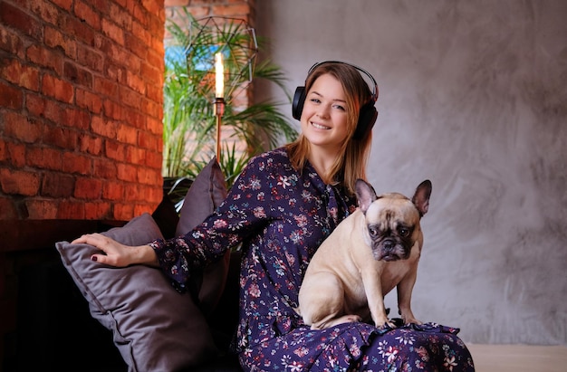 Happy blonde woman in dress sitting with her cute pug on a handmade sofa and listening to music in room with loft interior.