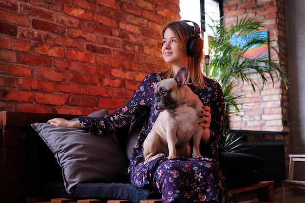 Happy blonde woman in dress sitting with her cute pug on a handmade sofa and listening to music in room with loft interior.