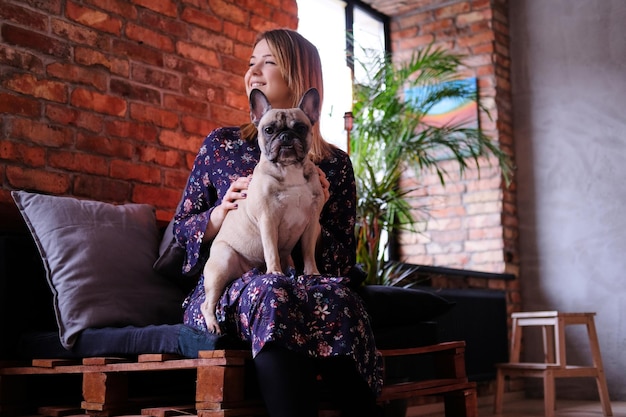 Happy blonde woman in dress sitting with her cute pug on a handmade sofa in a room with loft interior.