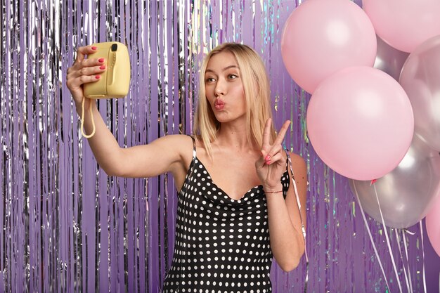 Happy blonde woman has makeup, keeps lips folded, makes selfie with small camera, stands against decorated wall with balloons and tinsel, wears stylish dress, shows peace hand gesture.