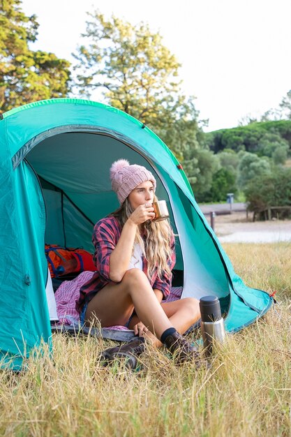 Happy blonde woman in hat drinking tea, sitting in tent and looking away. Caucasian long-haired traveler camping on lawn in park and relaxing on nature. Tourism, adventure and summer vacation concept