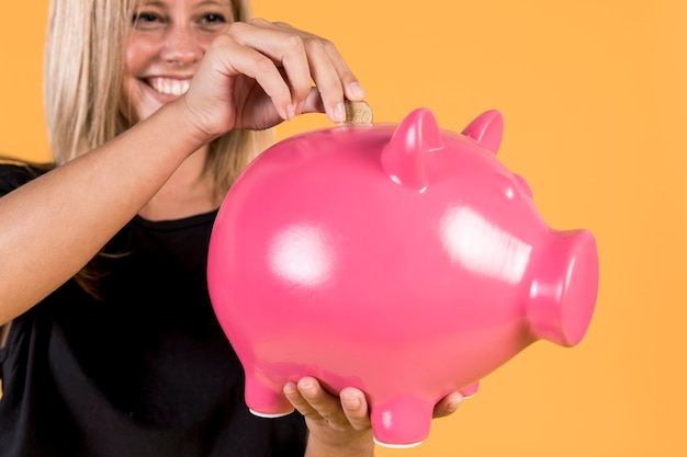 Donna bionda felice che inserisce moneta dentro il porcellino salvadanaio rosa