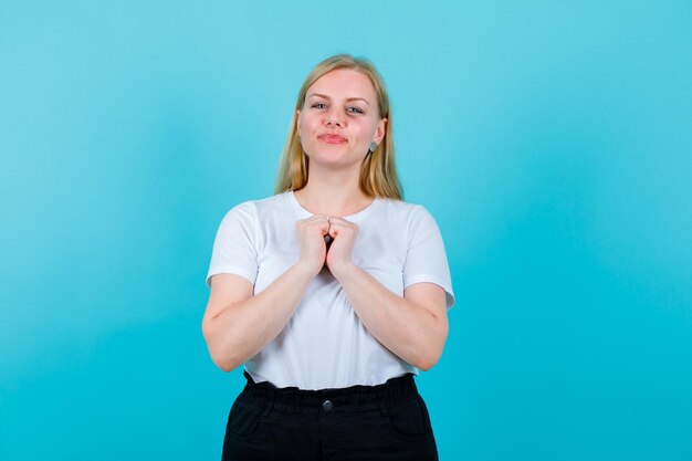 Happy blonde woman is looking at camera by holding fists on chest on blue background
