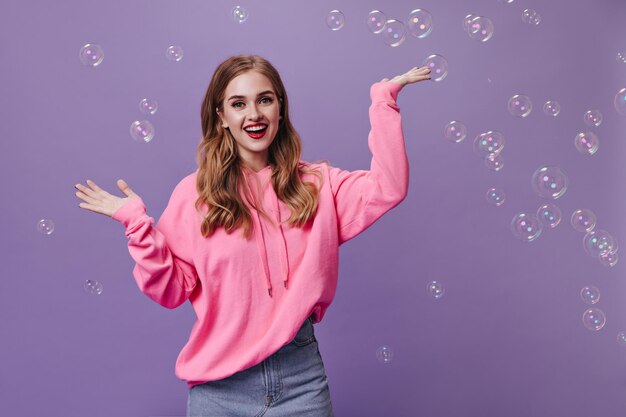 Happy blonde woman in pink sweatshirt playing with bubbles on purple background Young curly girl in denim skirt widely smiling on isolated