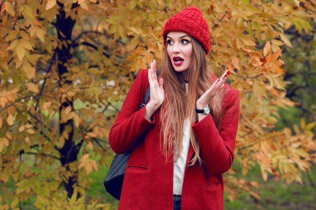 Счастливая белокурая женщина в красной шляпе и куртка представляя в парке осени.