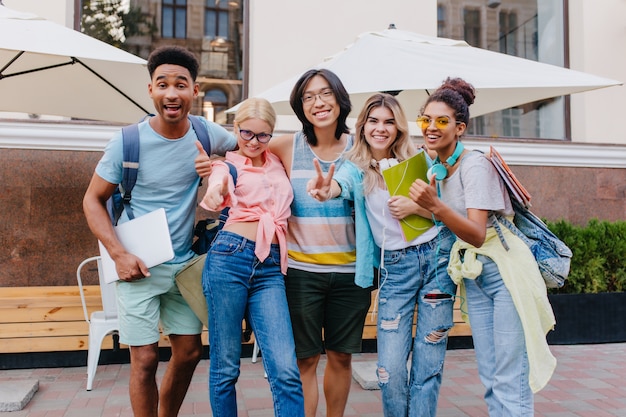 Happy blonde woman wears jeans with holes posing outdoor near smiling friends. Outdoor portrait of pleased students holding laptop and backpacks in morning.
