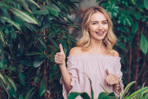 Счастливая белокурая молодая женщина стоя около зеленых растений показывая большой палец руки вверх по знаку