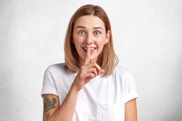 Happy blue eyed female with stylish haircut shows silence sign, asks not to spread information about her pregnancy, wants to make surpirsment for husband