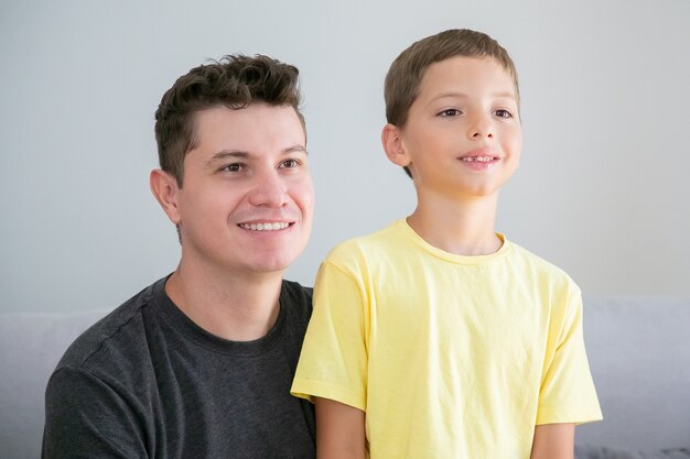幸せな少年と彼のお父さんは一緒に屋内に座って目をそらしています。ミディアムショット。家族と親の概念