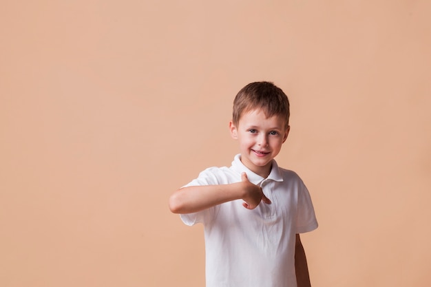 Счастливый мальчик, указывая пальцем на себя, стоя возле бежевой стены