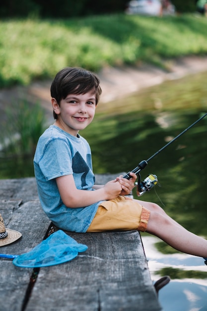 Happy boy sitting on wooden pier over lake holding fishing rod