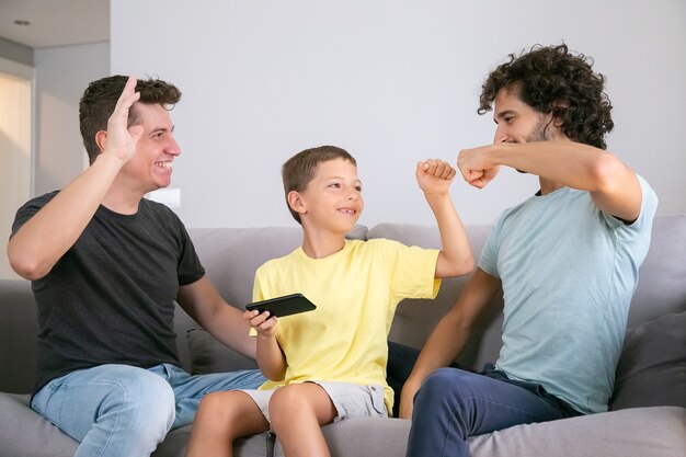 Happy boy with cellphone making fist bumping gesture with two cheerful dads. Fathers and son playing game on mobile phone together. Family at home and gay parents concept