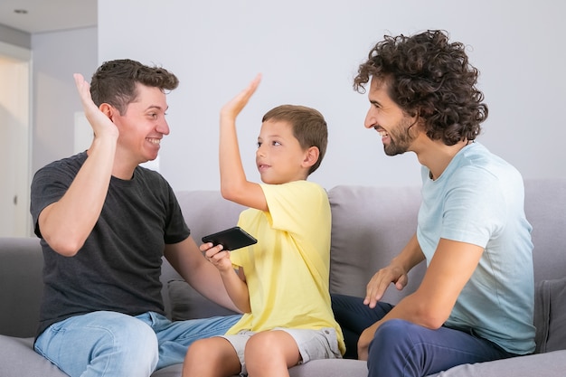 Happy boy with smartphone giving high five to two cheerful dads. fathers and son playing game on mobile phone together. family at home and gay parents concept