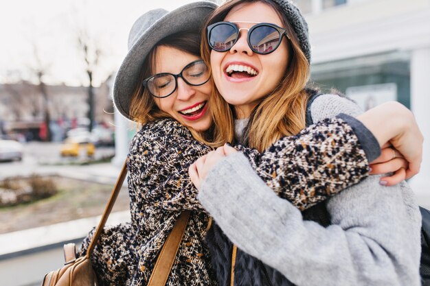 街の通りでハグする2人のスタイリッシュな女性の幸せな明るくポジティブな瞬間。クローズアップの肖像画面白い楽しいうれしそうな若い女性が楽しんで、笑顔、素敵な瞬間、親友。