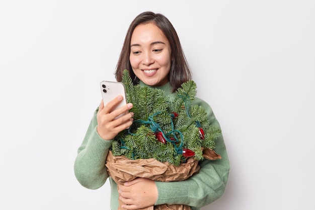 Счастливая брюнетка азиатская женщина счастливо смотрит на экран смартфона, получает поздравления в канун рождества, держит букет из еловых веток, позирует в помещении на фоне белой стены студии