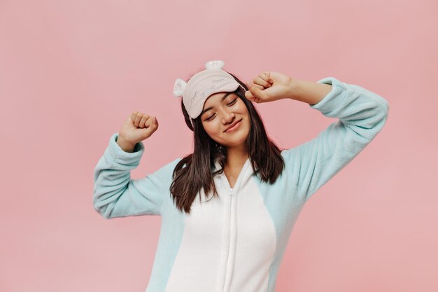 Happy brunette Asian woman in soft pajamas and sleep mask stretches and smiles on pink isolated background