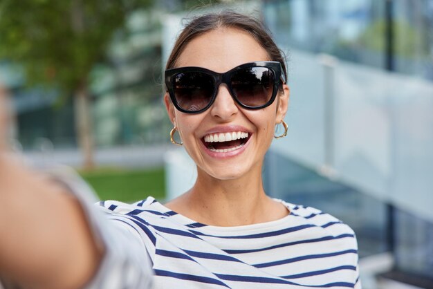 Happy brunette attractive woman wears trendy sunglasses and striped jumper keeps arm outstretched poses for selfie during leisure time at urban setting stands outdoors against blurred background