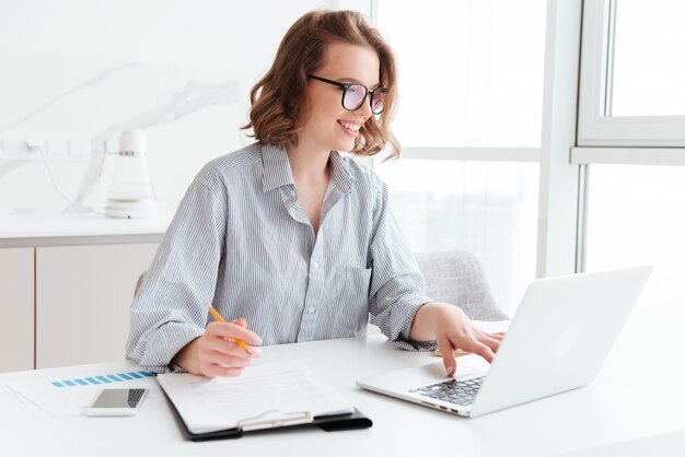 Happy brunette businesswoman in glasses using laptop computer while working in light apartment